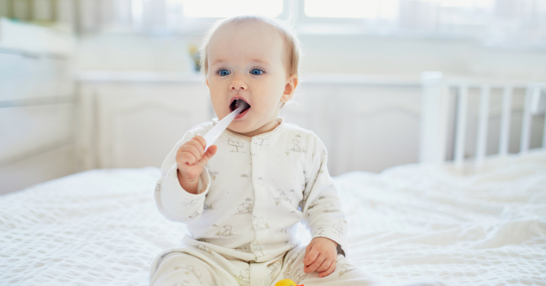 Baby boy sitting on bed with toothbrush in his mouth.