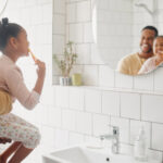 Father holds daughter while she brushes teeth in bathroom.