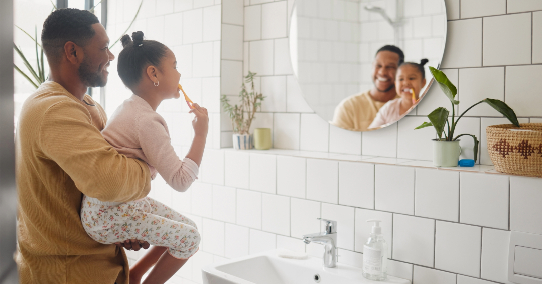 Father holds daughter while she brushes teeth in bathroom.