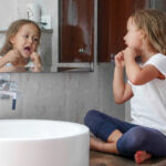 Young girl sitting on bathroom counter looking in mirror and flossing.