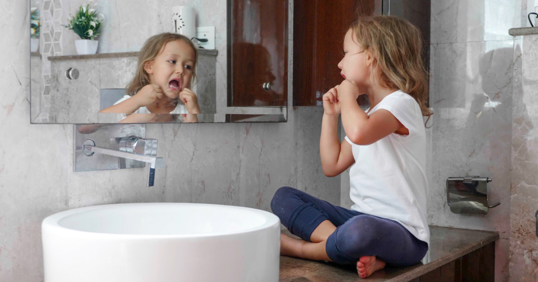 Young girl sitting on bathroom counter looking in mirror and flossing.