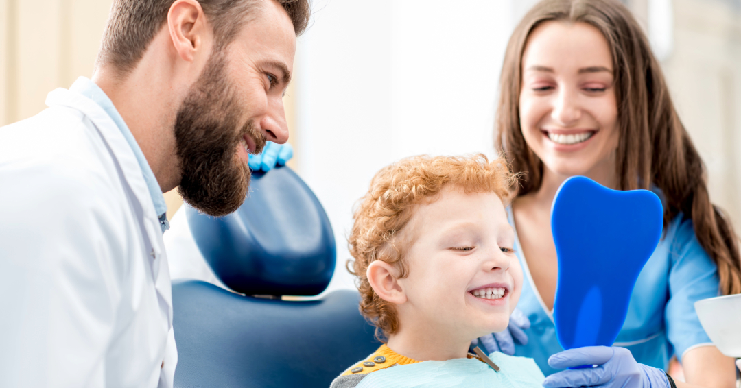 Pediatric dentist and dental assistant showing a child his smile in a tooth shaped mirror.