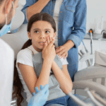 Girl sitting in dentist’s chair holding cheek.