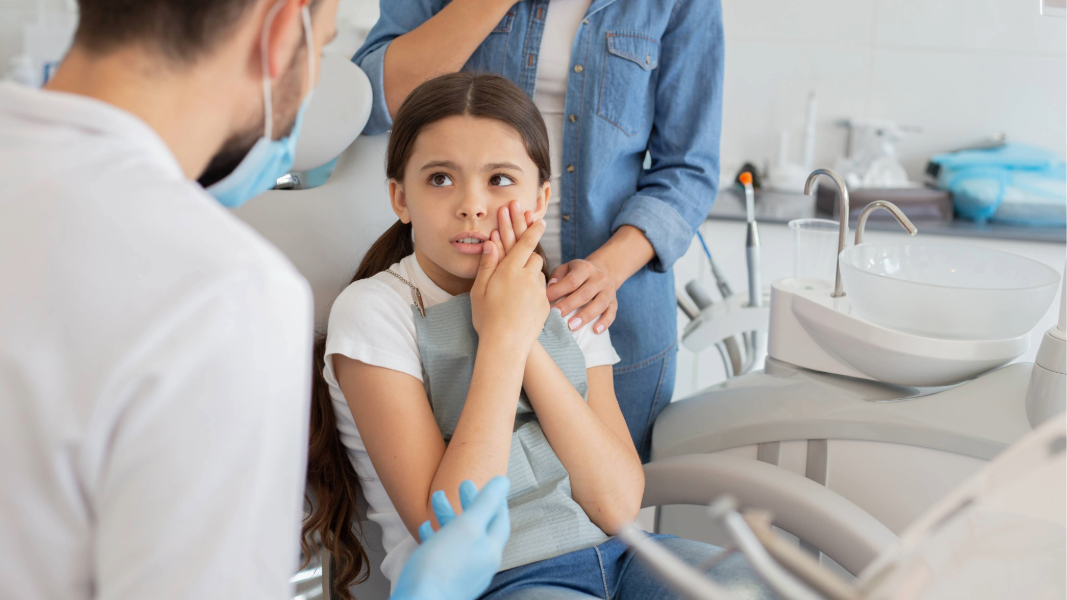 Girl sitting in dentist’s chair holding cheek.